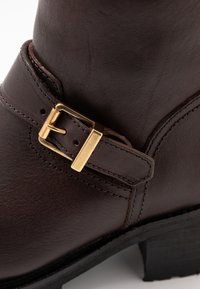 Hnědá kožená bota s texturovaným povrchem, zlatým kovovým přezkovým detailem a nízkým patem s viditelným šitím.