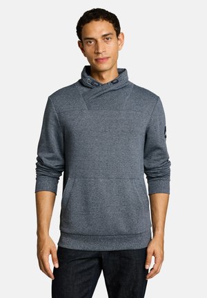 Mann mit kurzen lockigen Haaren, der einen grauen Pullover mit hohem Überkreuzkragen und dunkle Hosen trägt, steht vor weißem Hintergrund.