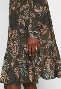 Robe vert foncé avec des motifs de feuilles roses, bleues et beiges, ourlet à volants et manchette boutonnée sur une main à la peau foncée portant des bagues en or.
