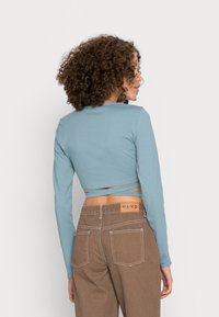 Top cropped de mangas longas em tecido azul suave com decote redondo. Combinado com calças castanhas de cintura alta com uma etiqueta e costura contrastante.