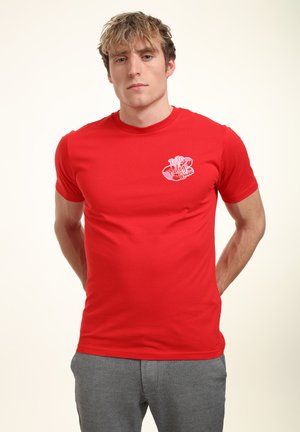 Giovane uomo che indossa una maglietta rossa con una piccola grafica bianca e rossa sul petto e pantaloni grigi, in piedi con le mani dietro la schiena.