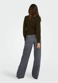 Femme aux cheveux bruns ondulés portant un pull vert foncé et un jean gris à taille haute et jambes larges, debout de dos à la caméra.