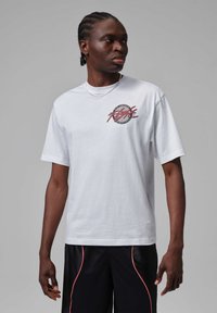 Hombre con cabello trenzado que lleva una camiseta blanca con diseño gráfico y pantalones cortos negros con ribete rojo, mirando hacia su izquierda contra un fondo liso.