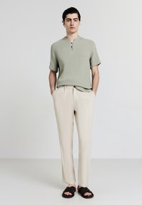 Camicia verde chiaro a maniche corte con una superficie testurizzata, dotata di colletto arrotondato e tre bottoni. Abbinata a pantaloni color crema e sandali neri.