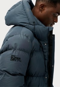 Chaqueta acolchada de color azul oscuro con capucha, que cuenta con un cierre de cremallera en el frente, cordón elástico para ajuste y un logo bordado en la manga.