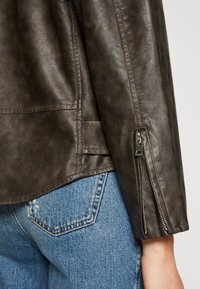 Veste en cuir marron foncé avec une texture lisse, comportant une fermeture éclair latérale sur la manche et une coupe ajustée sur un jean en denim bleu.