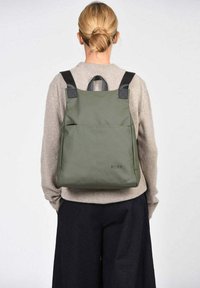 Mochila de tela verde con correas negras, forma rectangular, con un bolsillo frontal y un logotipo en la parte inferior.