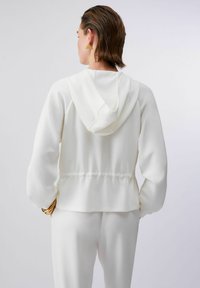 Femme aux cheveux bruns courts portant un sweat à capuche blanc et un pantalon assorti, accessoirisée avec des boucles d'oreilles triangles en or et des bracelets.