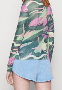 Langærmet, gennemsigtig bluse med en grøn bund og abstrakte pink- og hvide mønstre; parret med lyseblå shorts i en blød tekstur.