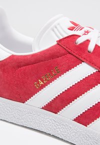 Červené semišové tenisky s bílou gumovou podrážkou, třemi bílými pruhy a zlatým logem "GAZELLE" na boku. Polstrovaný jazyk a kotníkový límec.