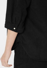 Close-up of a person's arm wearing a black corduroy shirt with a pearl button on the rolled-up sleeve cuff.