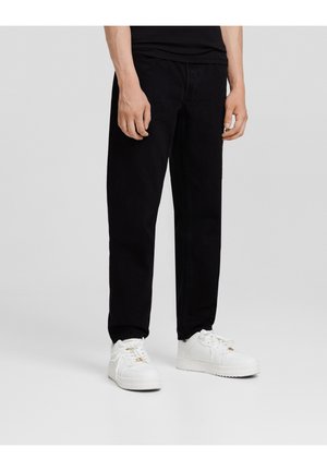 Homme portant un pantalon noir et des baskets blanches, debout devant un fond clair uni.