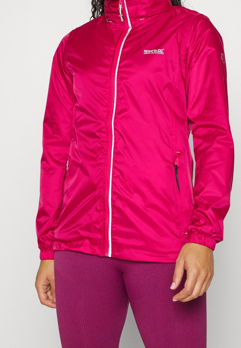 Femme portant une veste d'extérieur zippée rose vif avec des poignets élastiques et un logo sur la poitrine, associée à des leggings rose foncé.