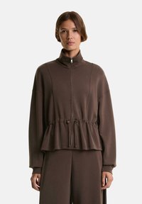 Brauner Kapuzenpullover mit hohem Kragen, gerafftem Saum, langen Ärmeln und einem weichen, strukturierten Stoff. Ausgestattet mit einem silbernen Reißverschluss und Kordelzügen.