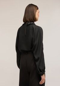 Blusa de satén negra con mangas abullonadas y cuello con tachuelas. La parte trasera presenta una abertura en forma de llave y una textura suave, combinada con pantalones negros a medida.