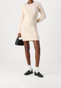 Robe-pull en tricot beige clair avec motifs torsadés et formes texturées en losange. Associée à un sac à main noir et des mocassins portés avec des chaussettes blanches.