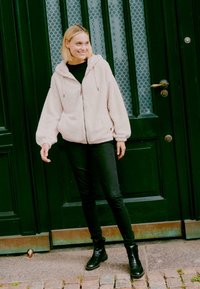 Heledad roosa fleece'ist zip-up jakk, millel on kapuuts ja elastsed mansetid, kantud musta topi üle ning koos kitsaste mustade teksade ja mustade saapatega.