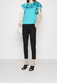 Blusa turquesa con mangas acampanadas, pantalones ajustados negros y sandalias de tacón bloque negro con punta abierta. La modelo sostiene un bolso blanco.