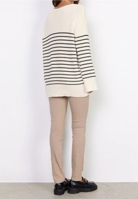 Cremefarbener Pullover mit schwarzen horizontalen Streifen, strukturierter Strick, lockere Passform, kombiniert mit beigen, taillierten Hosen und schwarzen Plateauschuhen.