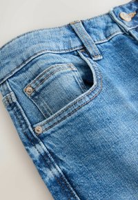 Détail d'un jean en denim bleu montrant la poche avant, la petite poche à monnaie, la passants de ceinture et le bouton métallique avec surpiqûres visibles.