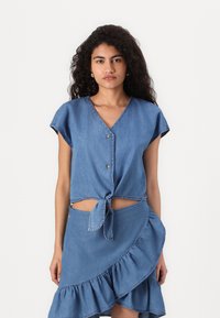 Lyseblå denimtopp med knutdetalj foran, kortermer og knappelukking. Kombinert med et matchende volanginnpakket skjørt.