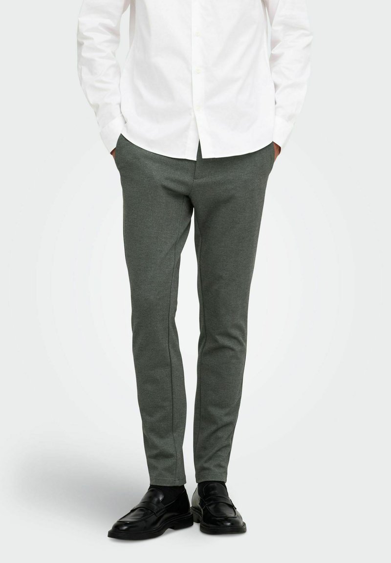 Άνδρας που φοράει σκούρο γκρι παντελόνι slim-fit, μαύρα loafers και λευκό πουκάμισο με κουμπιά, με τα χέρια στις τσέπες.