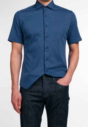 Mann trägt ein blaues Kurzarm-Hemd, das leicht in dunkle Jeans gesteckt ist, mit einer Hand in der Tasche vor weißem Hintergrund.