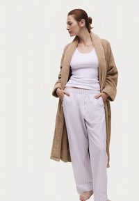 Beige lang kåbe med tekstur, hvid tanktop og lyse pyjamabukser med striber. Bukserne har knapper og sidelommer. Modellen står barfodet.