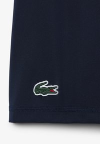 Camiseta polo azul marino con textura suave, que presenta un logo de cocodrilo verde con borde blanco cerca del dobladillo. Diseño sencillo y clásico.