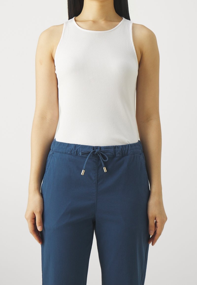 Weißes geripptes Tanktop mit breiten Trägern, kombiniert mit blauen Jogginghosen mit Kordelzug und goldfarbener Hardware. Glatte Textur, taillierte Passform.