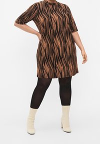 Sukienka z krótkim rękawem w brązowym kolorze z czarnym wzorem w zygzak, sięgająca do kolan, noszona na czarnych rajstopach i kremowych botkach za kostkę. Tkanina ma fakturowany połysk.