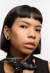 Base neutra media con SPF 50, llevada por un modelo. Presenta una bufanda negra con un patrón blanco, cabello corto oscuro y textura de piel suave.