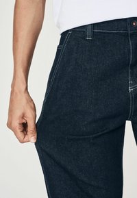 Donkerblauwe denim broek met contrasterende witte stiksels en een knoopsluiting. De stof is stevig en heeft een rechte beenontwerp.