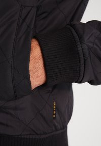 Hand in die Tasche einer schwarzen, gesteppten Jacke mit geripptem Bund und kleinem goldenen "GABBA 1973"-Label gesteckt.