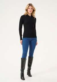 Black ribbed long-sleeve top with a zip collar, paired with blue fitted jeans and knee-high black boots. Textured knit material throughout.