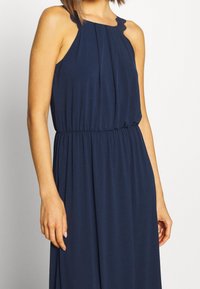 Granatowa, bezrękawnikowa sukienka z plisami w okolicy biustu, elastycznym pasem oraz delikatnym koronkowym wykończeniem na ramionach, wykonana z miękkiego materiału.
