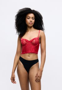 Bustier in pizzo rosso e raso con coppe con stecche e orlo scallopato, abbinato a uno string thong nero a vita alta. Texture liscia e design aderente.