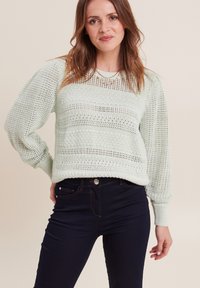 Hellmintgrüner gestrickter Pullover mit einem strukturierten Muster und gerippten Bündchen, kombiniert mit dunkelblauen Jeans.