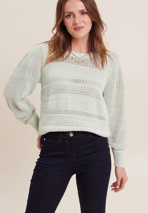 Hellmintgrüner gestrickter Pullover mit einem strukturierten Muster und gerippten Bündchen, kombiniert mit dunkelblauen Jeans.