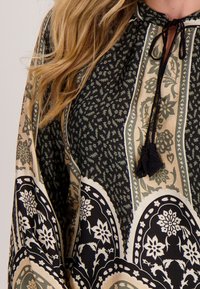 Vloeiende blouse met een zwart, crèmekleurig en olijfgroen bloemenpatroon, met een trekkoord bij de hals en uitgebreide ontwerpelementen.