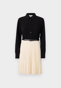 Haut noir à boutons avec des manches longues, associé à une jupe plissée crème. Présente une fine ceinture noire à la taille. Texture de tissu lisse.