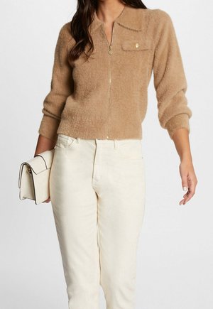 Femme portant un pull zippé beige duveteux avec une poche poitrine, un pantalon crème taille haute, tenant un sac à main beige clair dans sa main gauche.