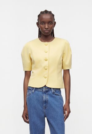 Γυναίκα με πλεξούδες, που φοράει μια χρυσαφί κοντομάνικη ζακέτα με κουμπιά και μπλε ψηλόμεσα τζιν μπροστά σε απλό φόντο.