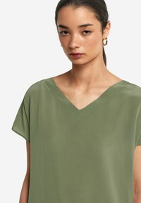 Olivgrön, kortärmad topp med V-ringning; jämn textur och avslappnad passform. Har en diskret sömdetalj längs halsringningen.