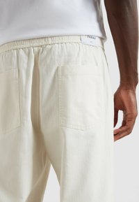 Jasno beżowe spodnie o fakturze w jodełkę, z elastycznym pasem i dwoma tylnymi kieszeniami. Metka z logo "FARAH" na pasie.