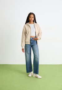 Bež zip-up dukserica, sivi crop top, traperice visoki struk u plavoj boji, bijele tenisice s ružičastim detaljima, stoji na zelenom tepihu.