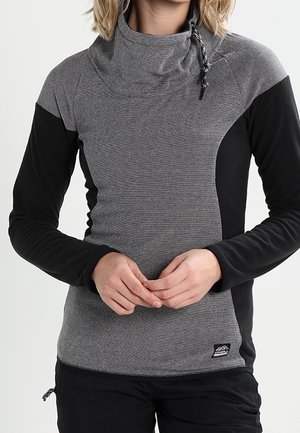 Kobieta w dwukolorowym, szaro-czarnym swetrze z długim rękawem, z wysokim kołnierzem i sznurkiem, ręce złożone na talii.