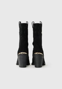 Bottes de cowboy mi-mollet en daim noir avec des talons empilés, des coutures décoratives et un détail métallique au-dessus du talon, présentées de l'arrière sur un fond blanc.