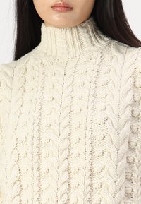 Pull turtleneck en chunky crème avec un motif en maille torsadée et des reliefs, accompagné d'un col côtelé. Tissu texturé et confortable.