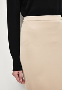 Beige kne-lång kjol med en texturerad yta, hög midja och rena sömmar, ihopklädd med en figurnära svart långärmad topp där ärmarna syns.